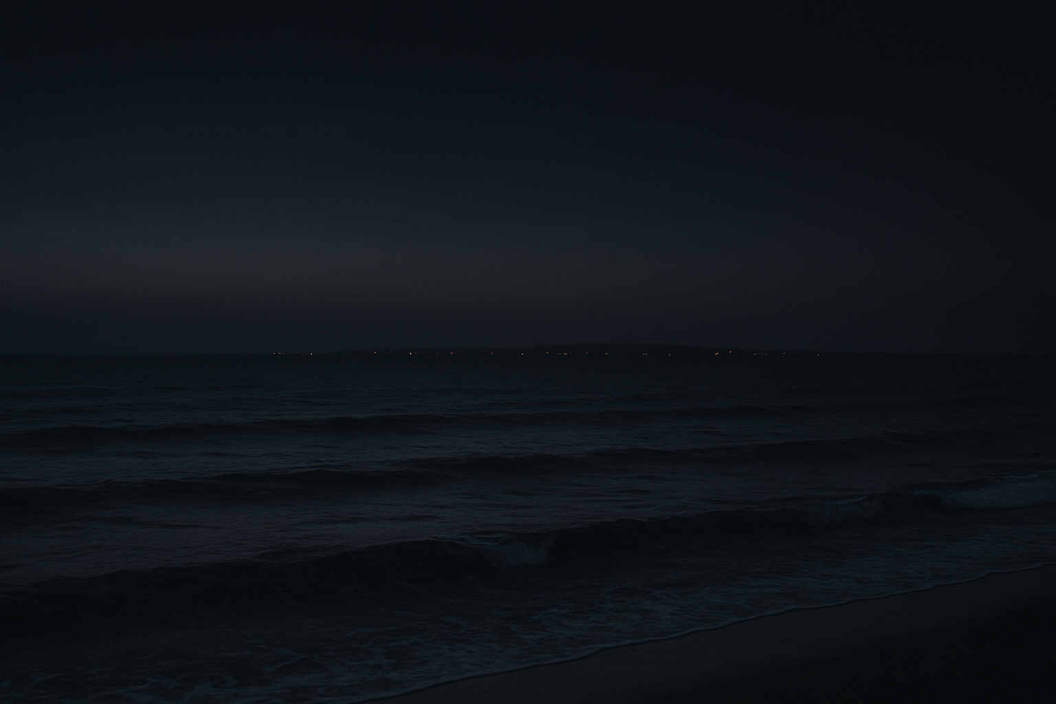 Dark coastal horizon at dusk with gentle waves and a faint line of lights in the distance