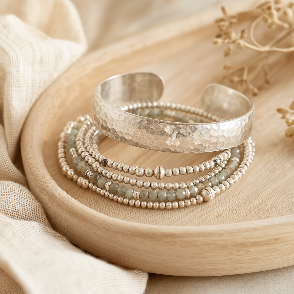 Hand-hammered silver cuff bracelet stacked with thin beaded bracelets on a wooden tray