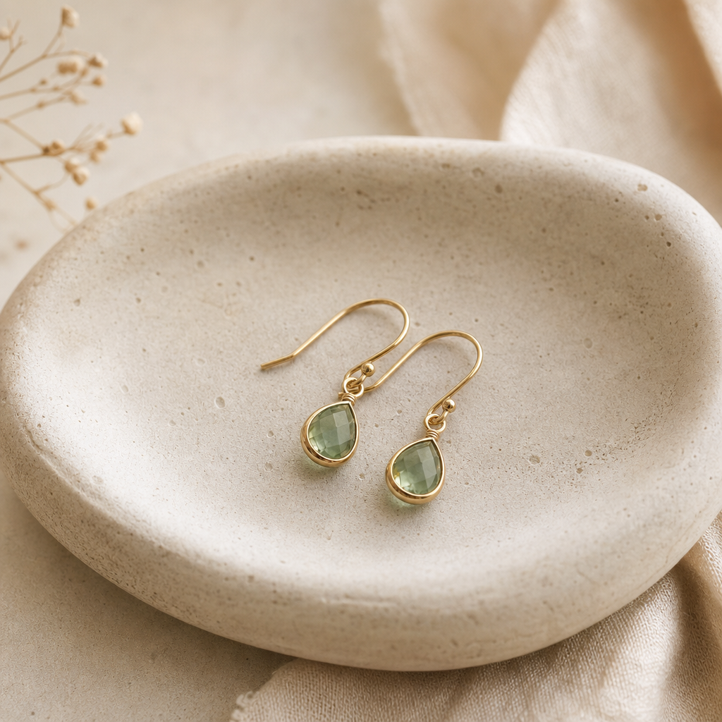 Pair of small sage-green gemstone drop earrings resting on a rounded stone dish
