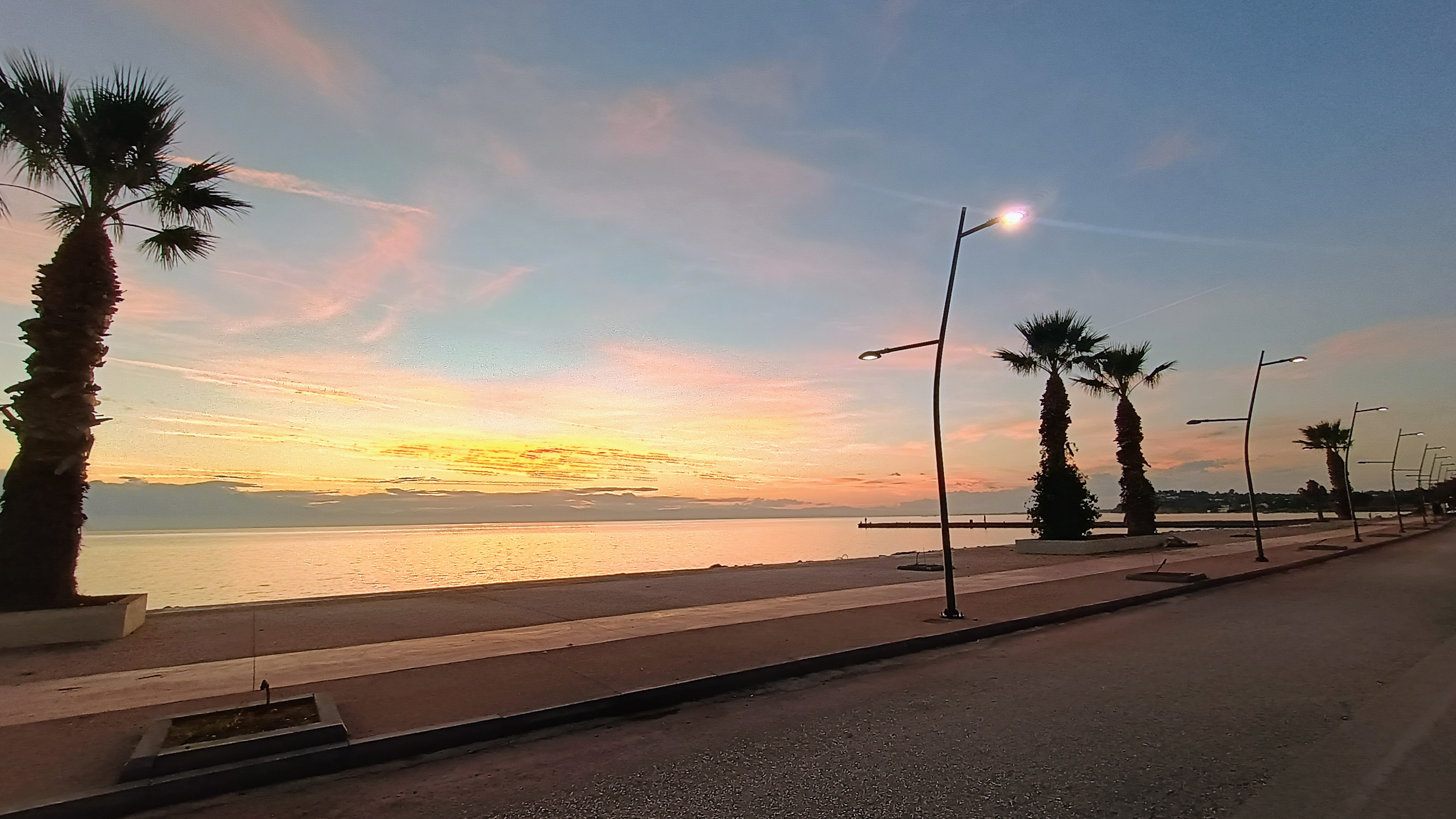 Seaside dusk promenade in Greece with palm-lined waterfront and streetlights