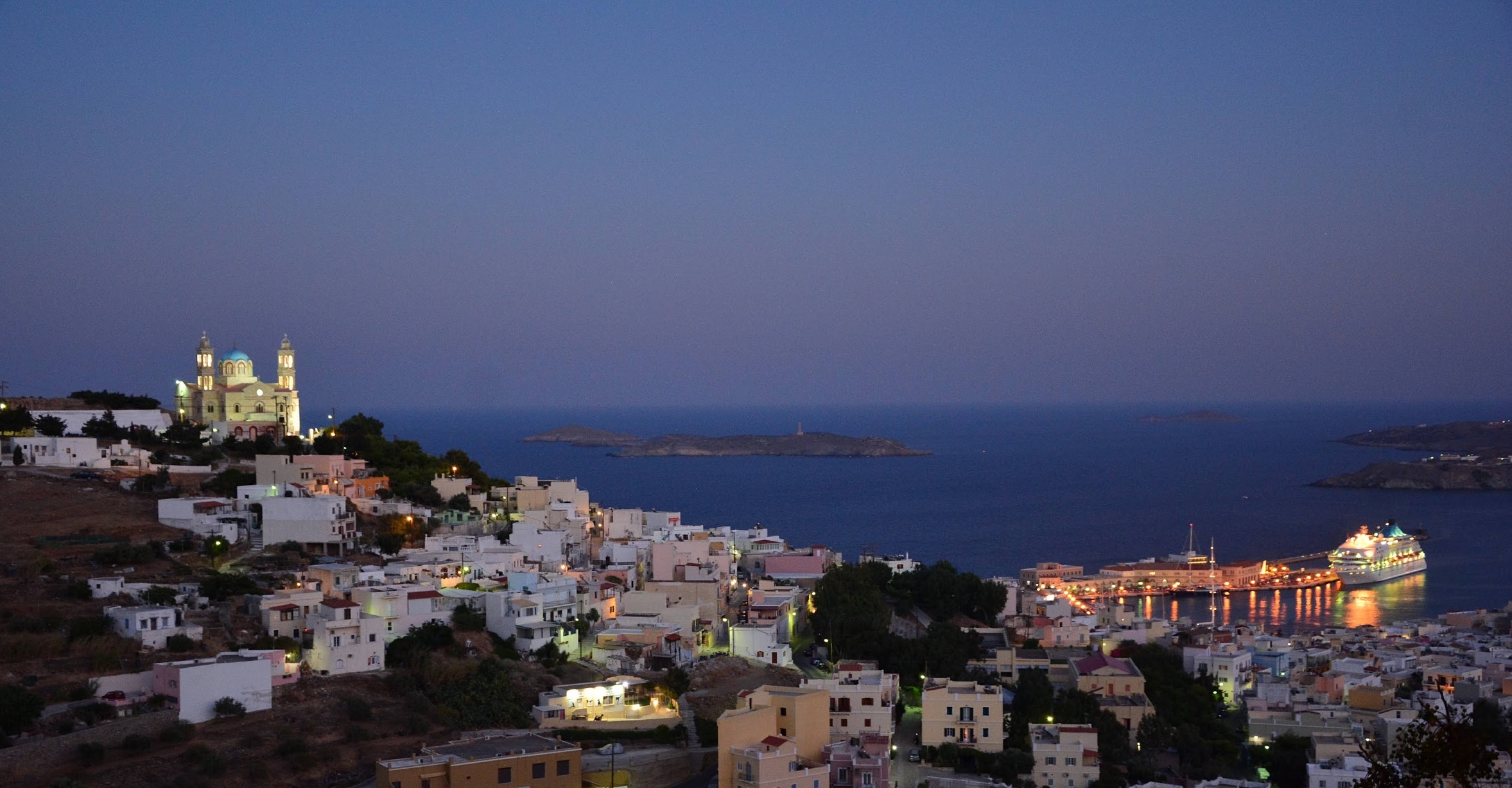 Dusk view over a Greek island harbor with hillside homes, a church on the ridge, and lit waterfront at blue hour