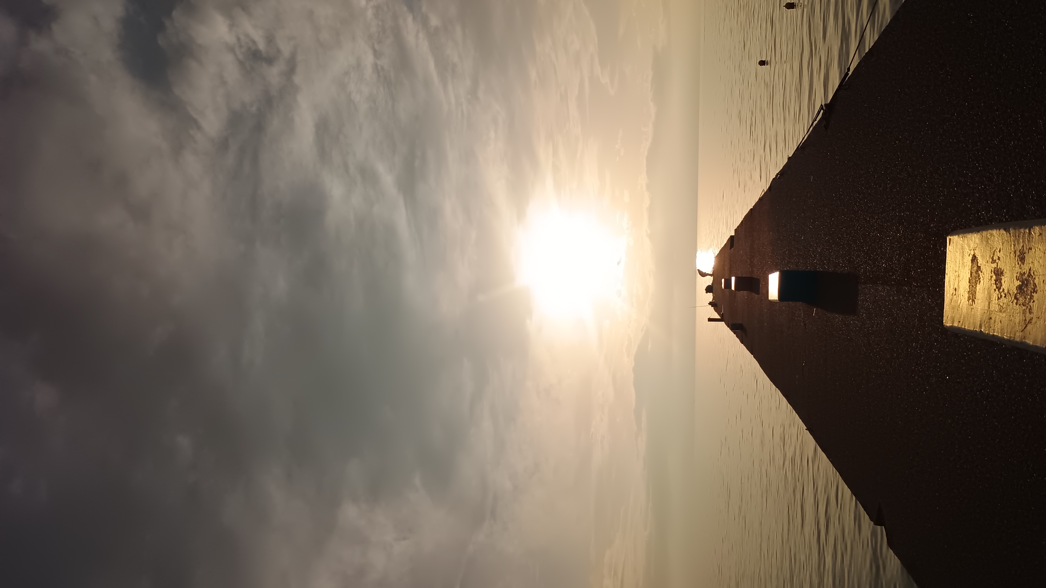 Seaside pier stretching toward a bright low sun under dramatic cloud cover, with a calm horizon line