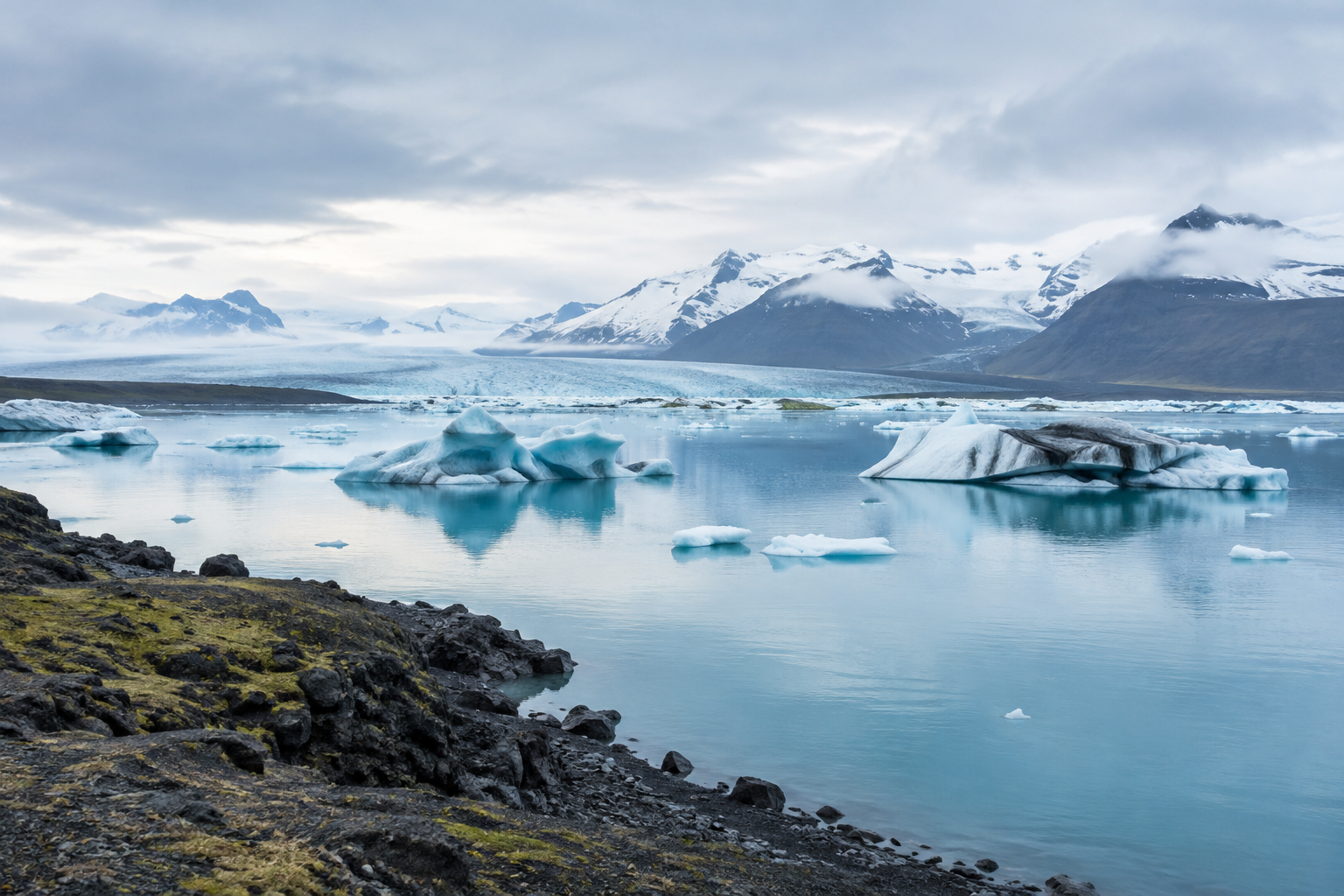A calm Icelandic glacial lagoon with pale blue water, dark volcanic shoreline, and distant ice-capped mountains beneath a soft overcast sky.