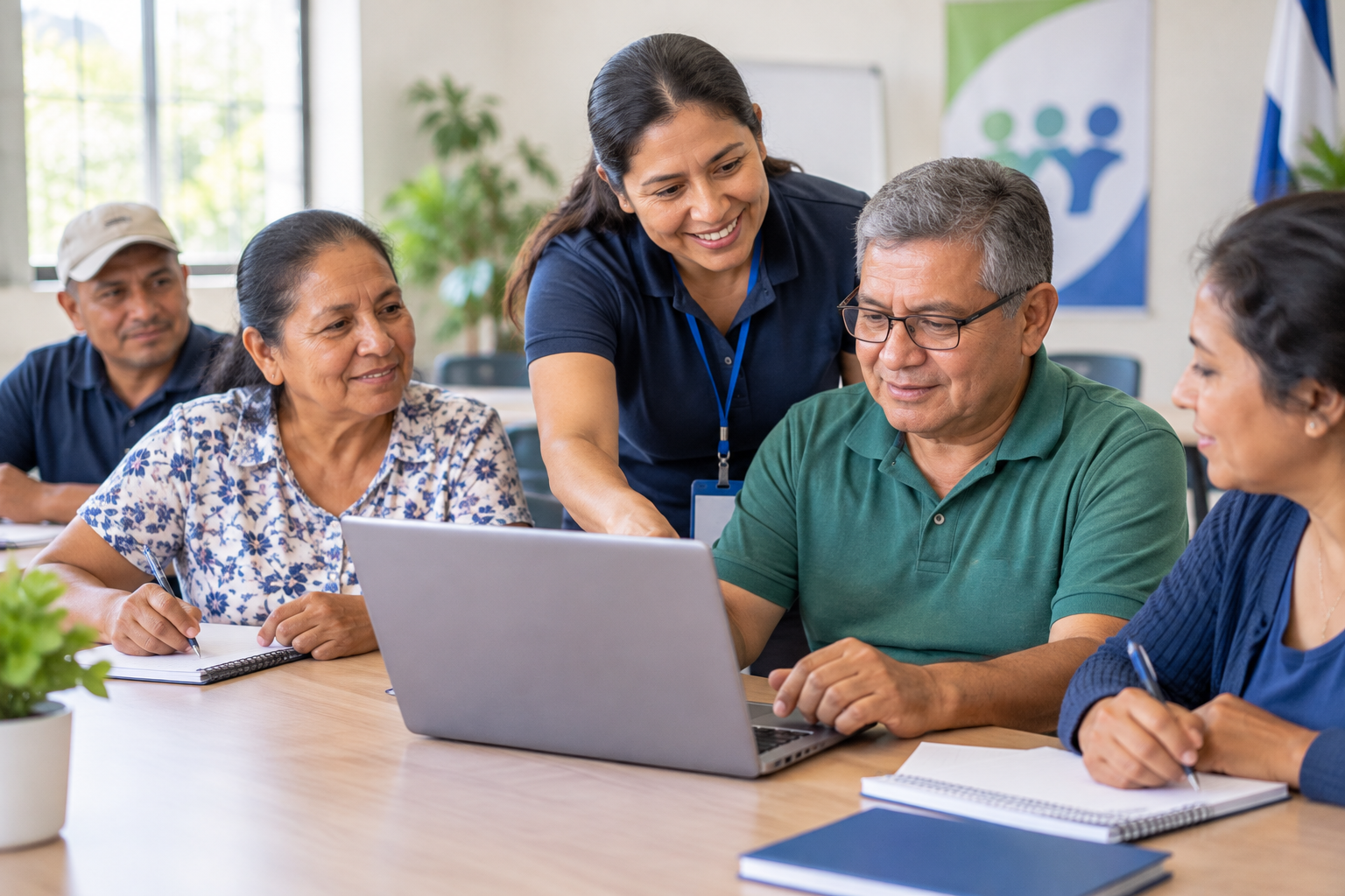 Facilitadora comunitaria acompañando a un pequeño grupo de personas adultas en una sesión de formación práctica con cuadernos y una computadora portátil