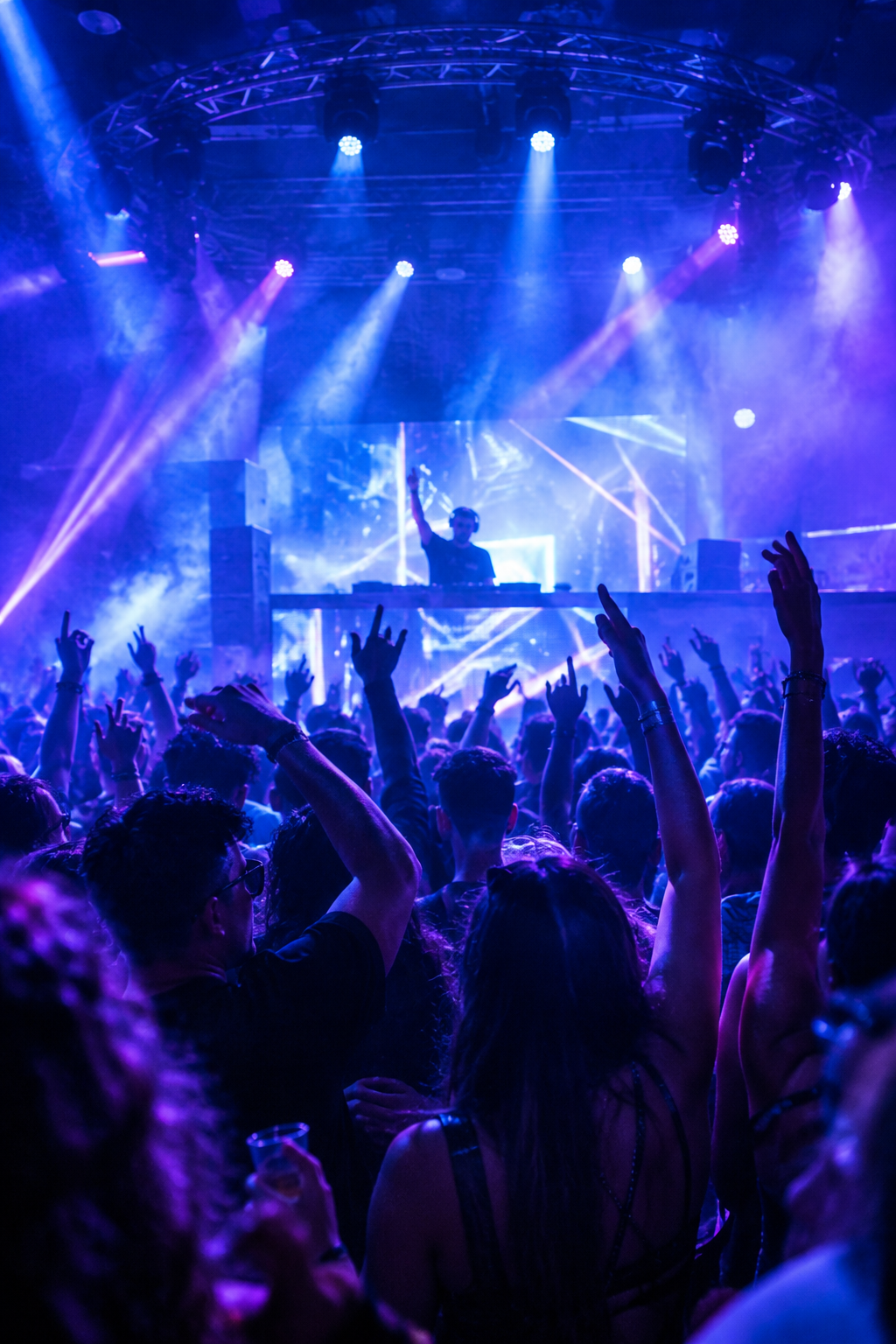 Packed nightlife crowd under cool blue and violet lights with raised hands and a DJ booth in front