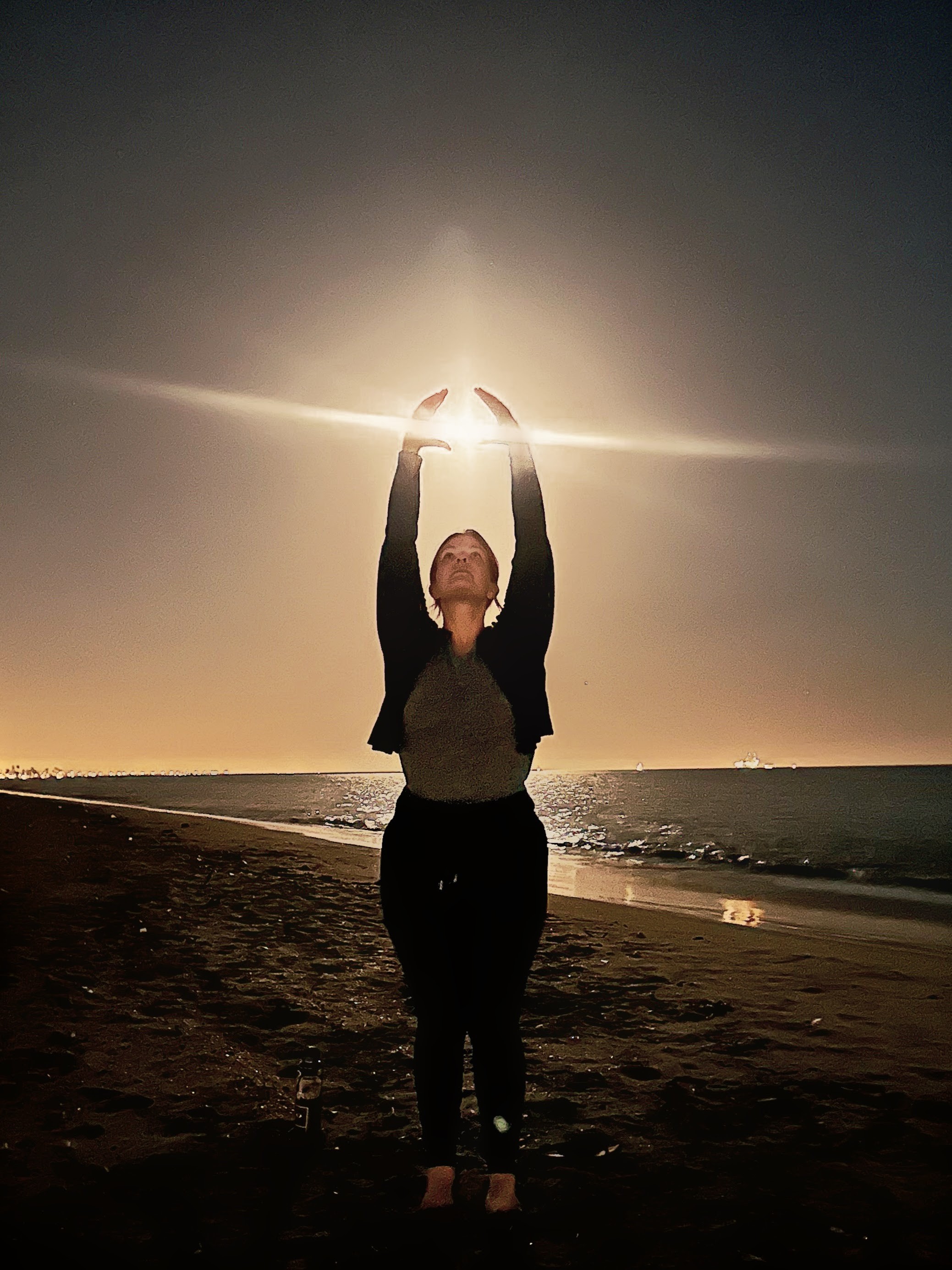 Silhouetted person on a beach at sunset with raised arms toward bright light over the ocean, creating a calm and hopeful wellness mood