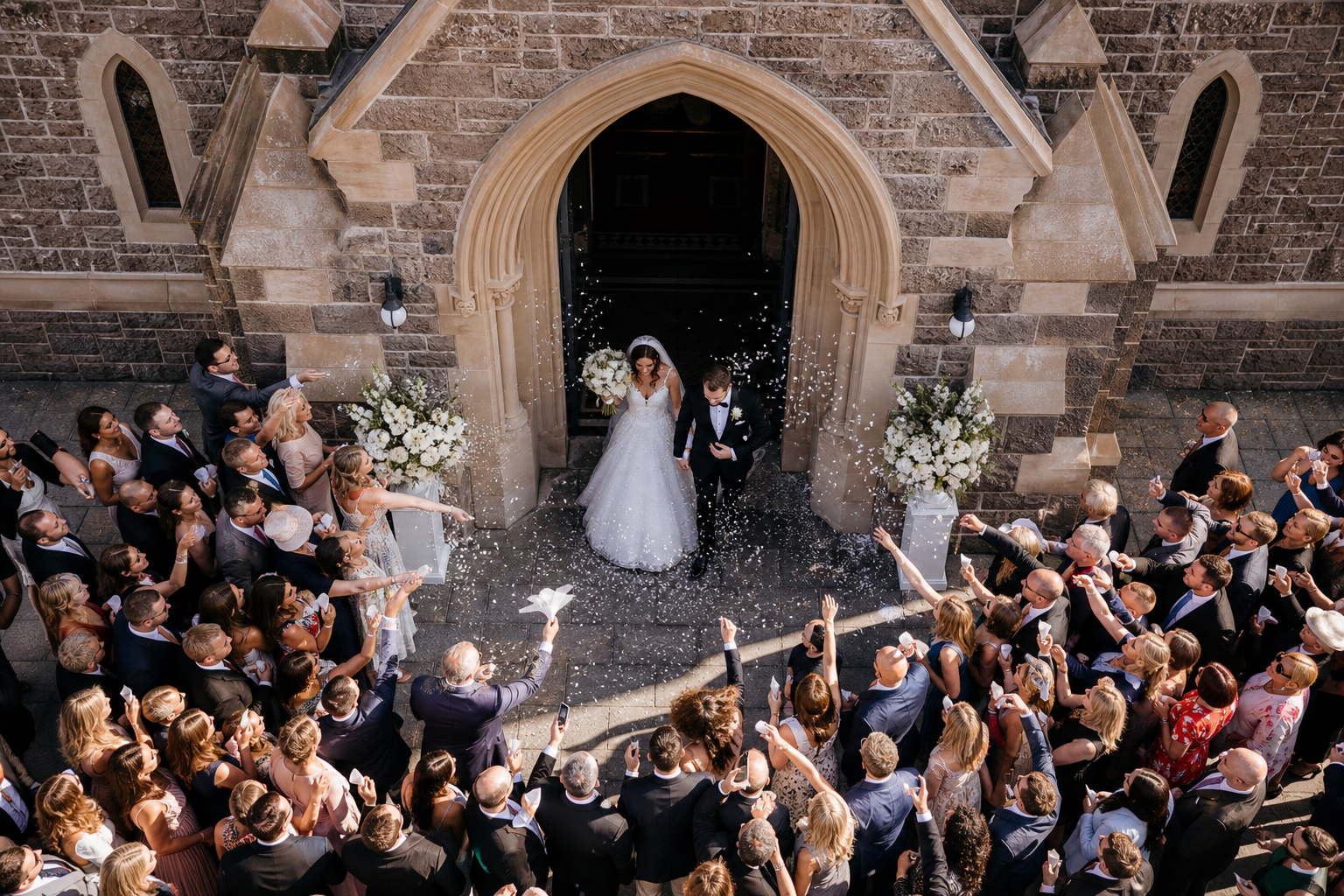 Drone frame of newlyweds exiting a stone church while guests throw white confetti