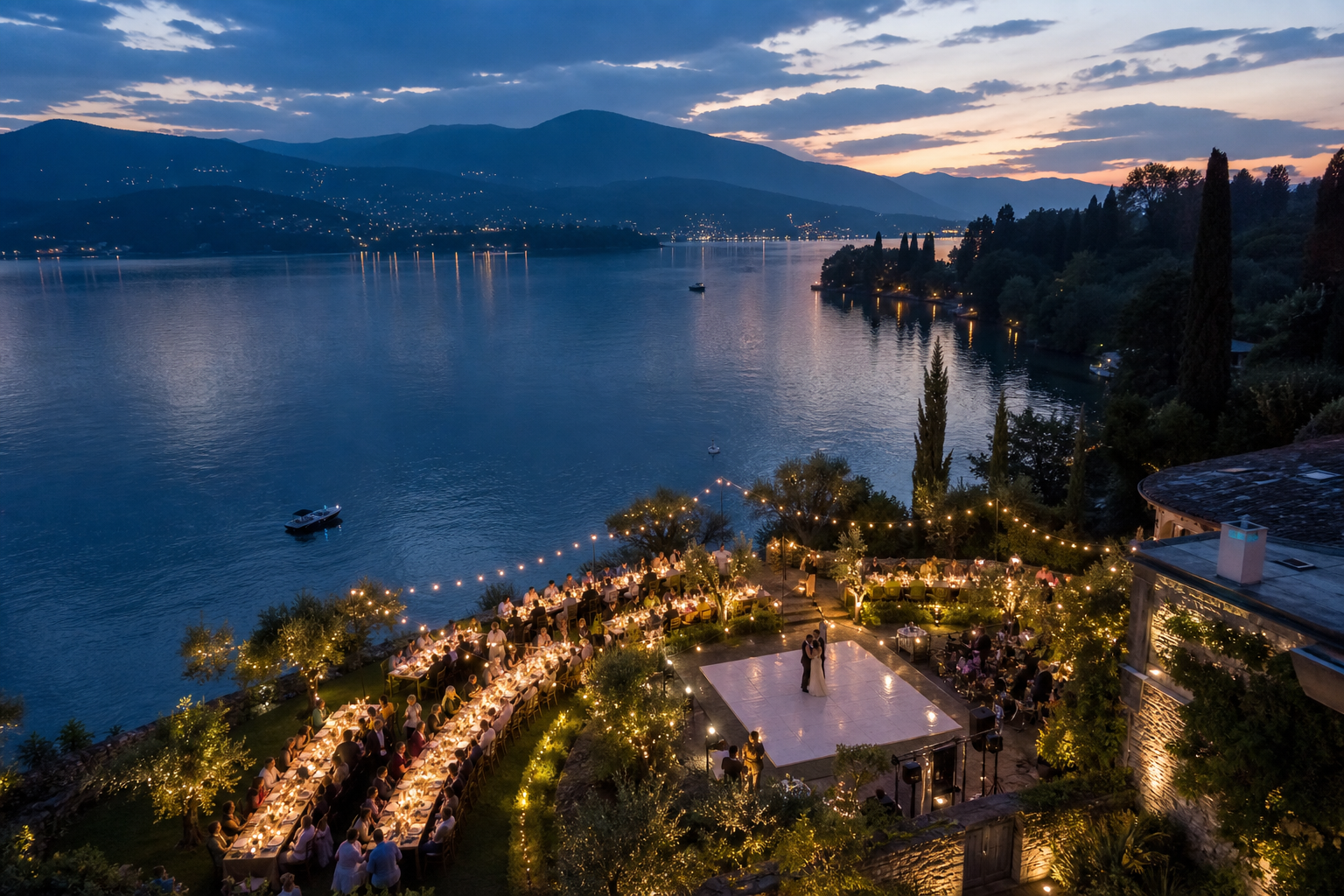 Aerial dusk view of a lakeside wedding reception with glowing string lights and long banquet tables