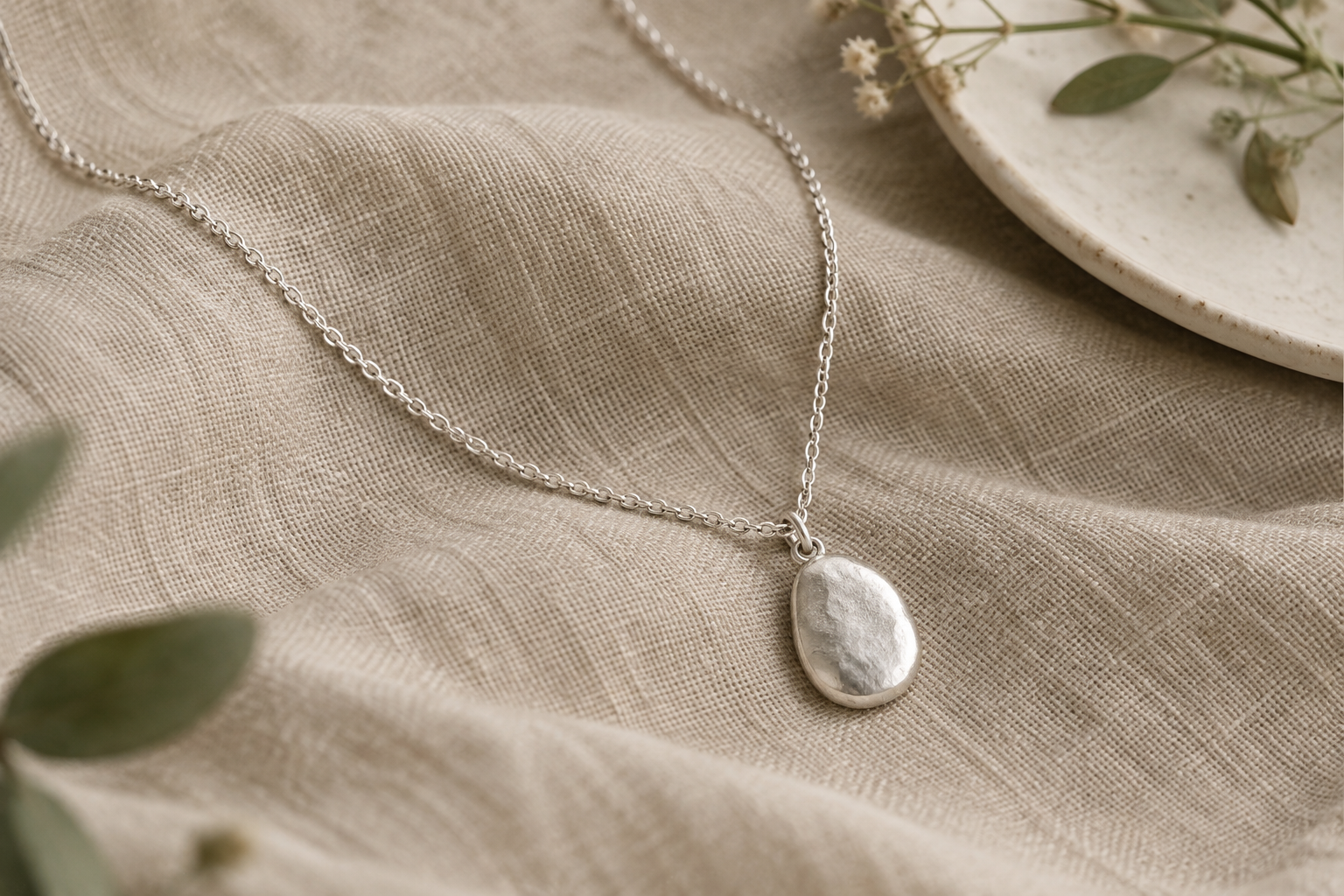 Fine sterling silver chain with an organic oval pebble pendant displayed on soft textured fabric