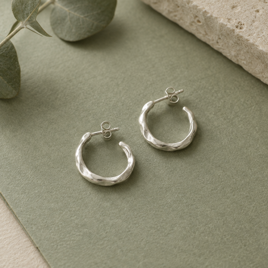 Pair of small hand-formed sterling silver hoop earrings with softly irregular contours on muted sage paper