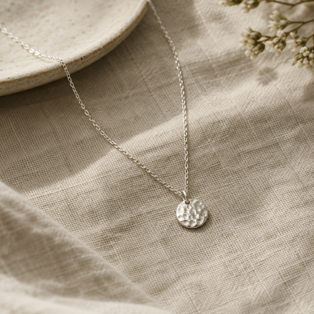 Delicate sterling silver necklace with a small hammered pendant arranged on natural flax linen