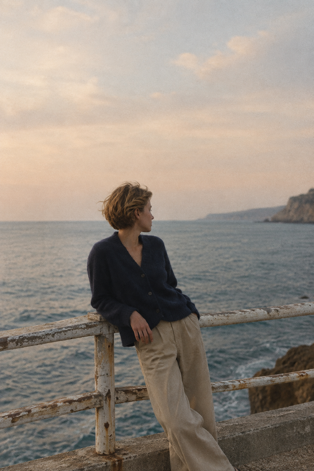 Figure in a navy cardigan beside a weathered seaside railing under a hazy pastel sky