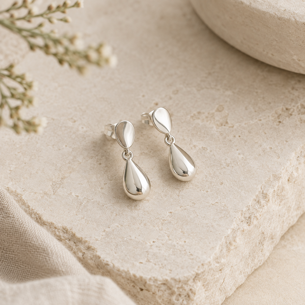 Pair of petite silver drop earrings arranged on a softly textured pale stone surface