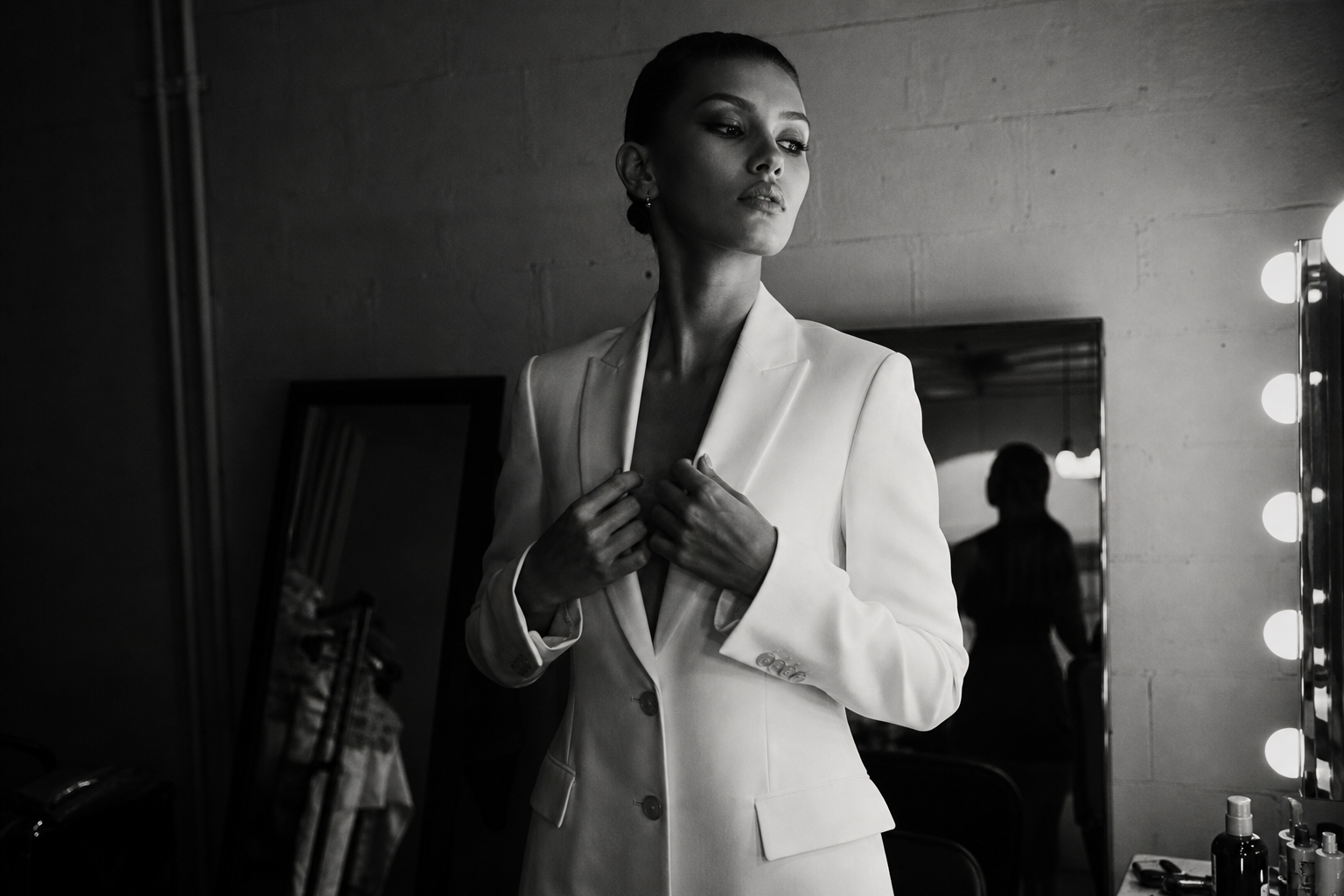 Backstage editorial moment in monochrome with a model adjusting a white blazer near industrial mirrors