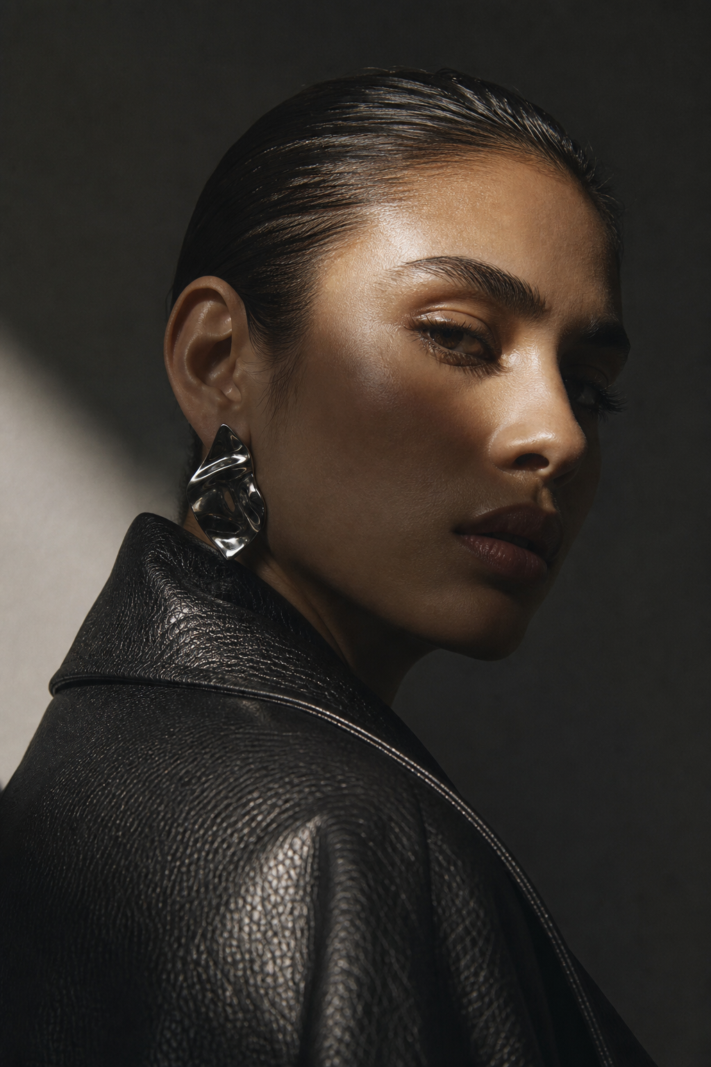 Close fashion portrait of a model with slick hair, silver earrings, and textured leather jacket under directional light