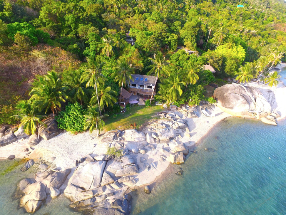 Aerial tropical shoreline at SOWK in Koh Phangan with palm-covered hillside, granite rocks, white sand, and clear turquoise water
