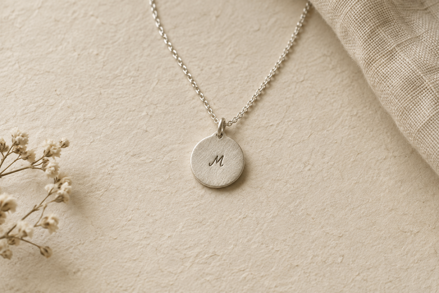 A brushed silver disc pendant engraved with a tiny handwritten initial on textured paper