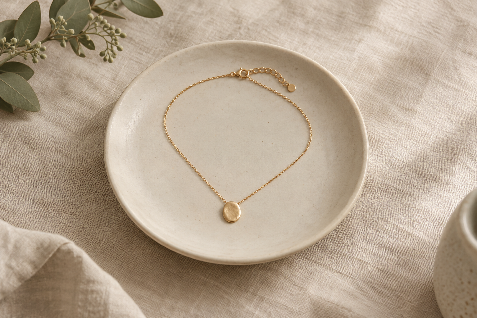 A fine gold necklace with an oval pendant resting on a matte ceramic dish