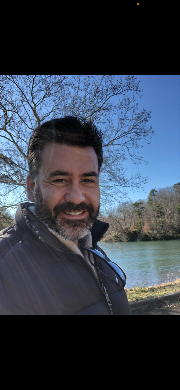 Lorenzo Cola smiling outdoors beside a calm lake under a clear blue sky