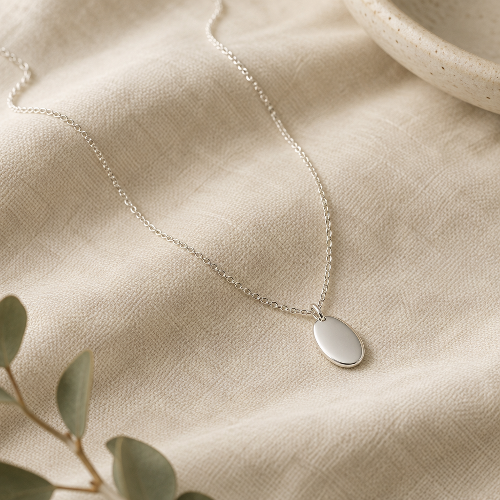 Delicate sterling silver necklace with a small oval pendant displayed on warm neutral fabric
