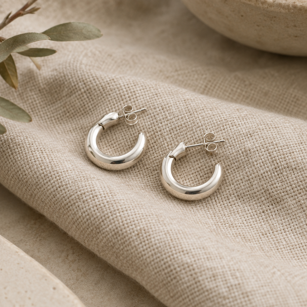 Pair of petite silver arc hoop earrings with a polished finish on oatmeal linen
