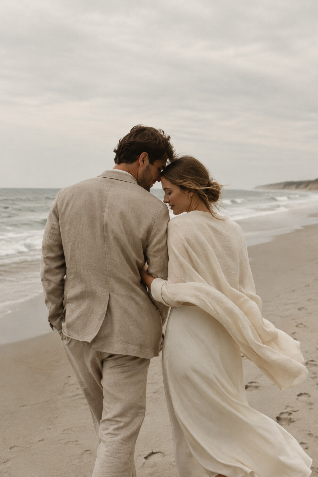 A couple in muted sand and cream clothing walk close together along a pale, windswept coastline beneath an overcast sky.
