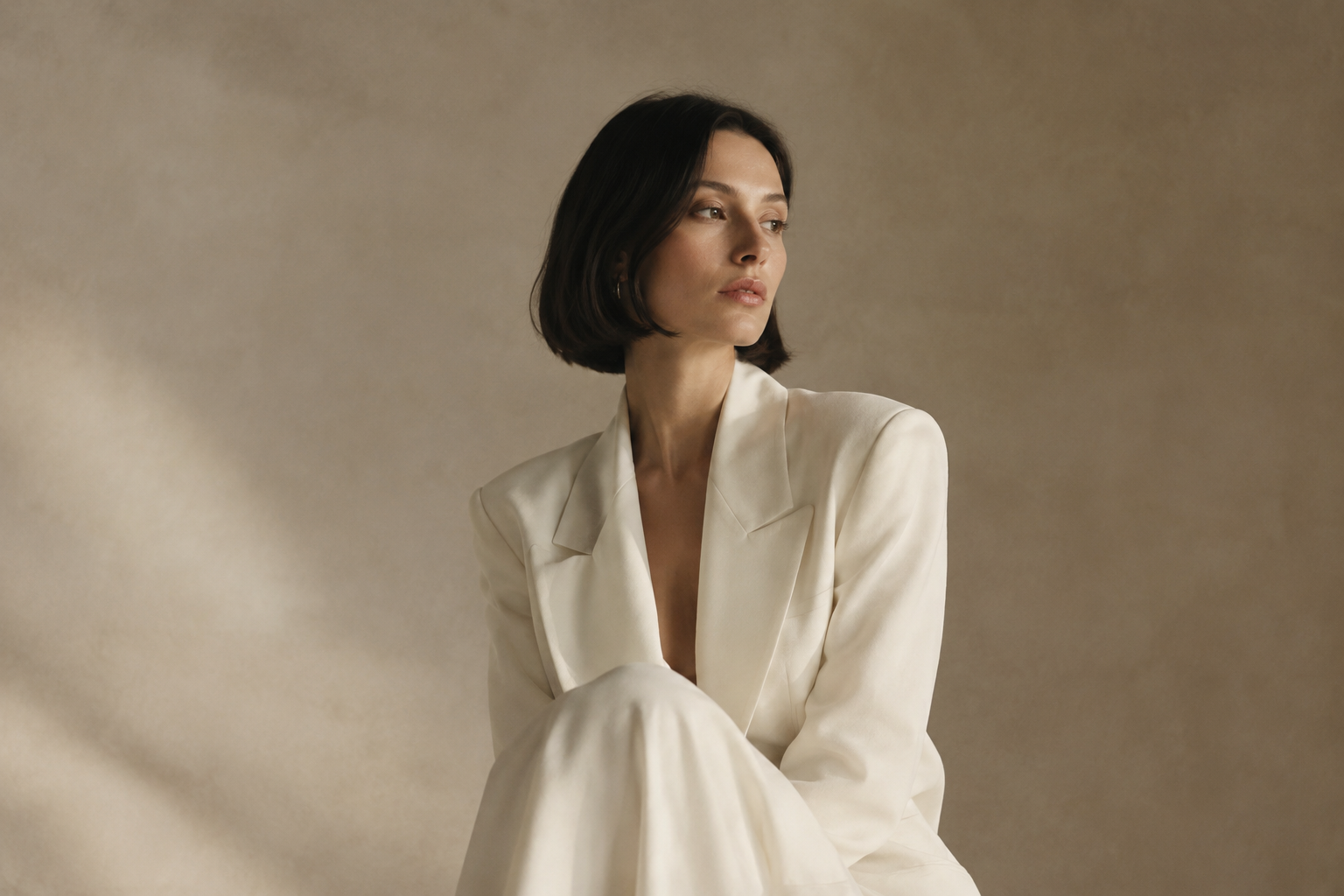 A poised woman with a sleek dark bob in a sculptural ivory outfit, photographed against a softly textured warm studio backdrop.