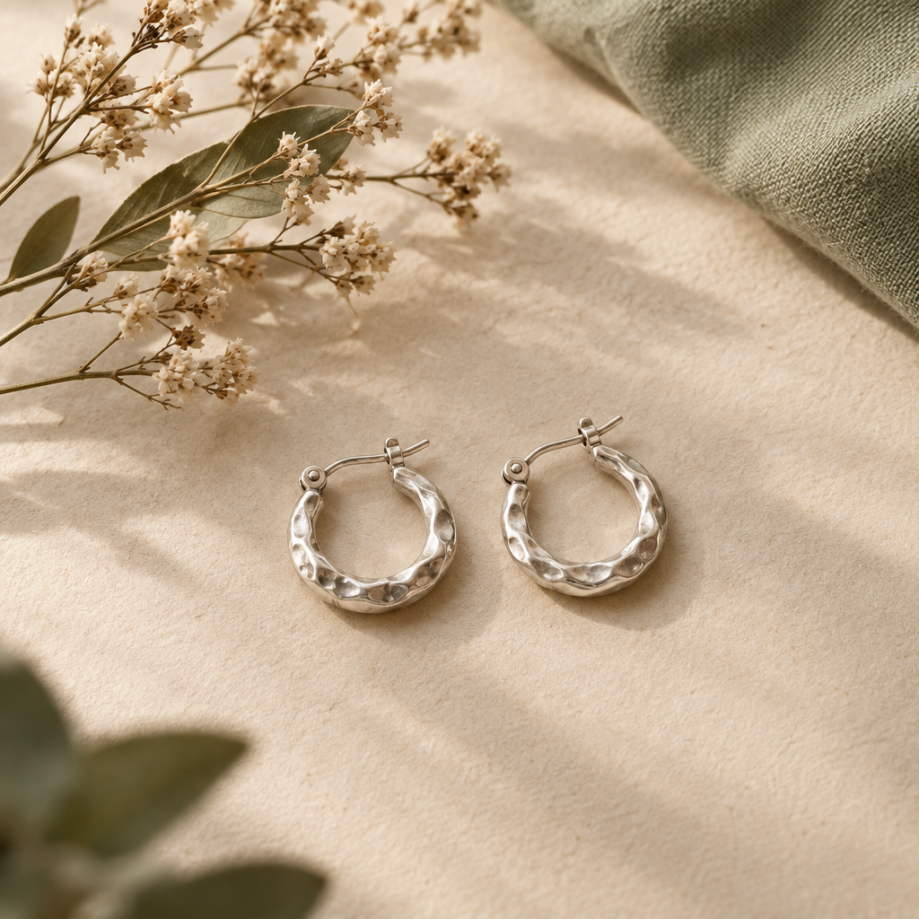 A pair of small hand-hammered silver hoop earrings beside dried botanical stems
