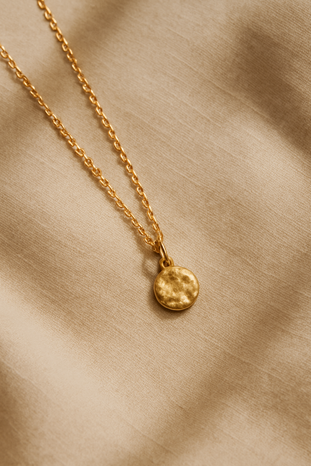 Delicate gold pendant necklace with a small textured charm laid on warm beige linen