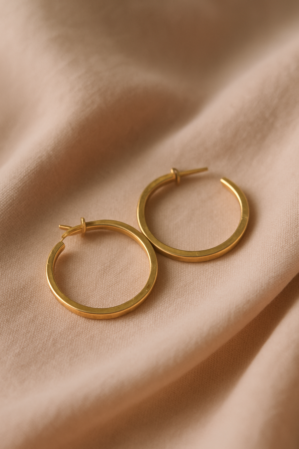 Pair of minimalist hammered gold hoop earrings on a soft blush fabric background