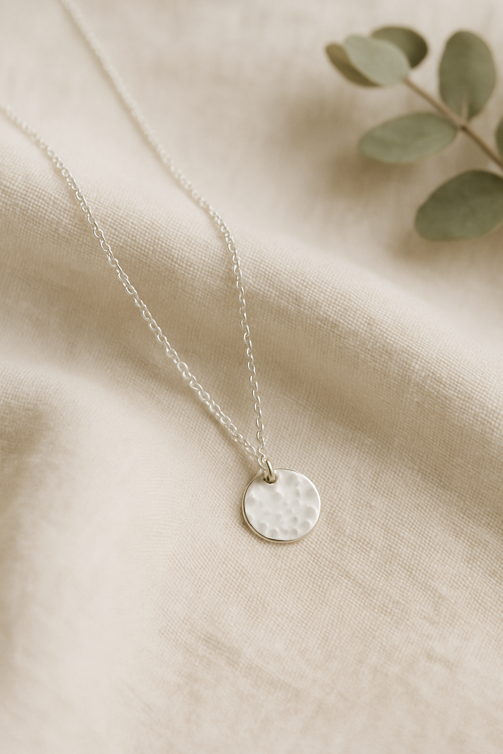 Delicate handmade sterling silver necklace with a small hammered disc pendant draped over soft cream linen