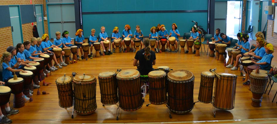 Indoor group drumming circle with participants seated around a wooden floor and multiple hand drums