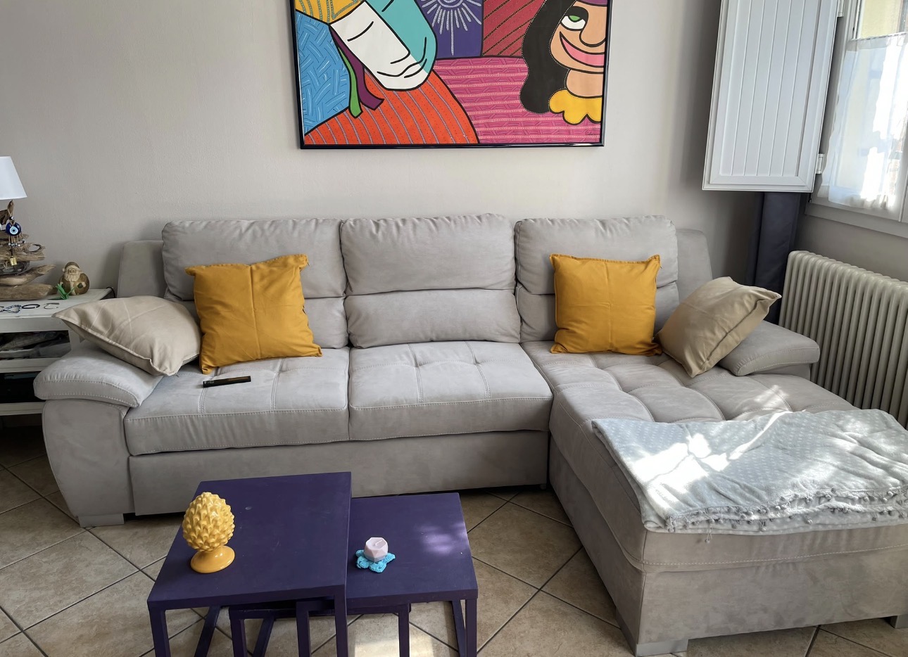 Soggiorno con divano angolare grigio chiaro, cuscini gialli, tavolini viola e quadro colorato sopra il divano