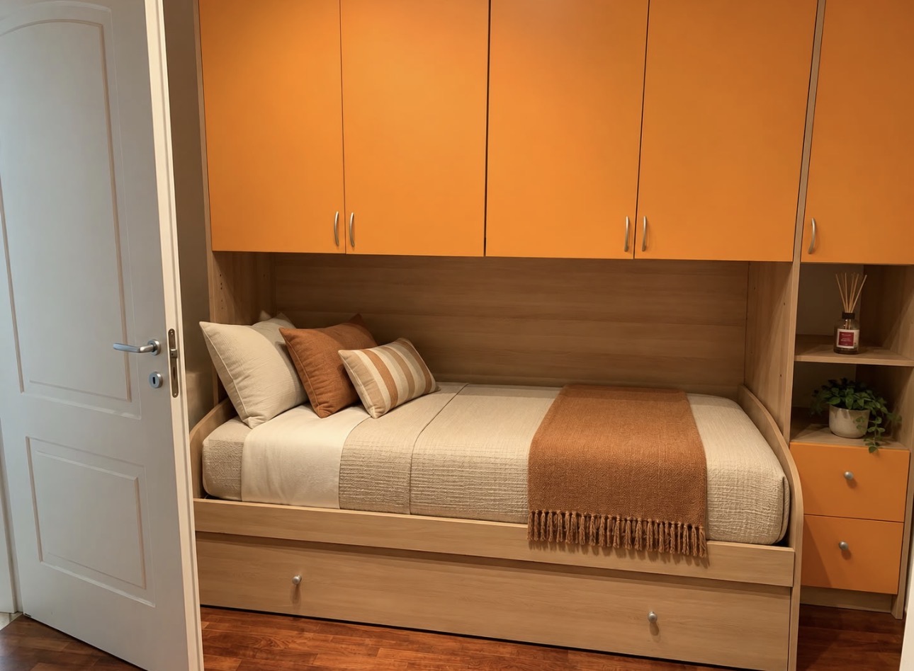 Camera con letto singolo incassato in struttura in legno chiaro, pensili arancioni e tessili nei toni caldi