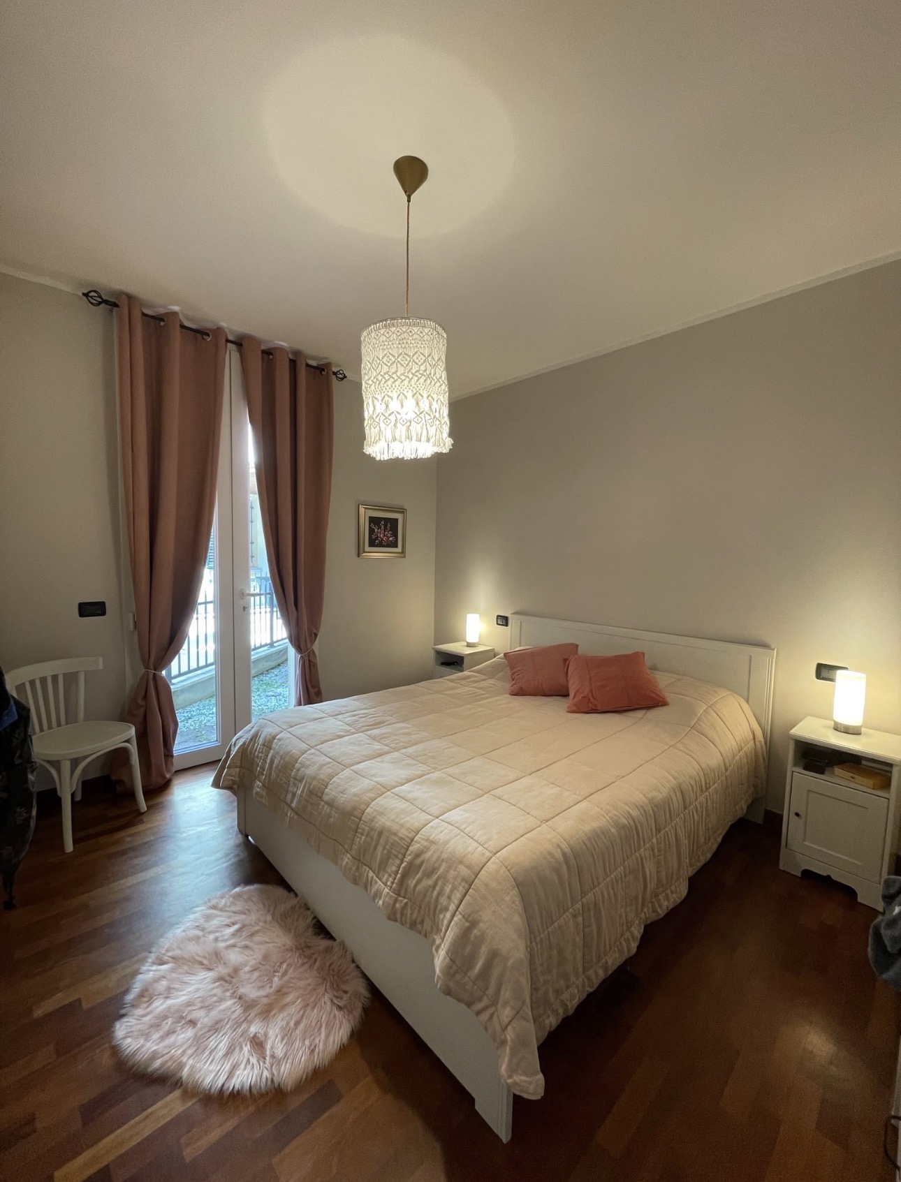 Camera matrimoniale luminosa con letto bianco, tende rosate, lampade accese e parquet in legno