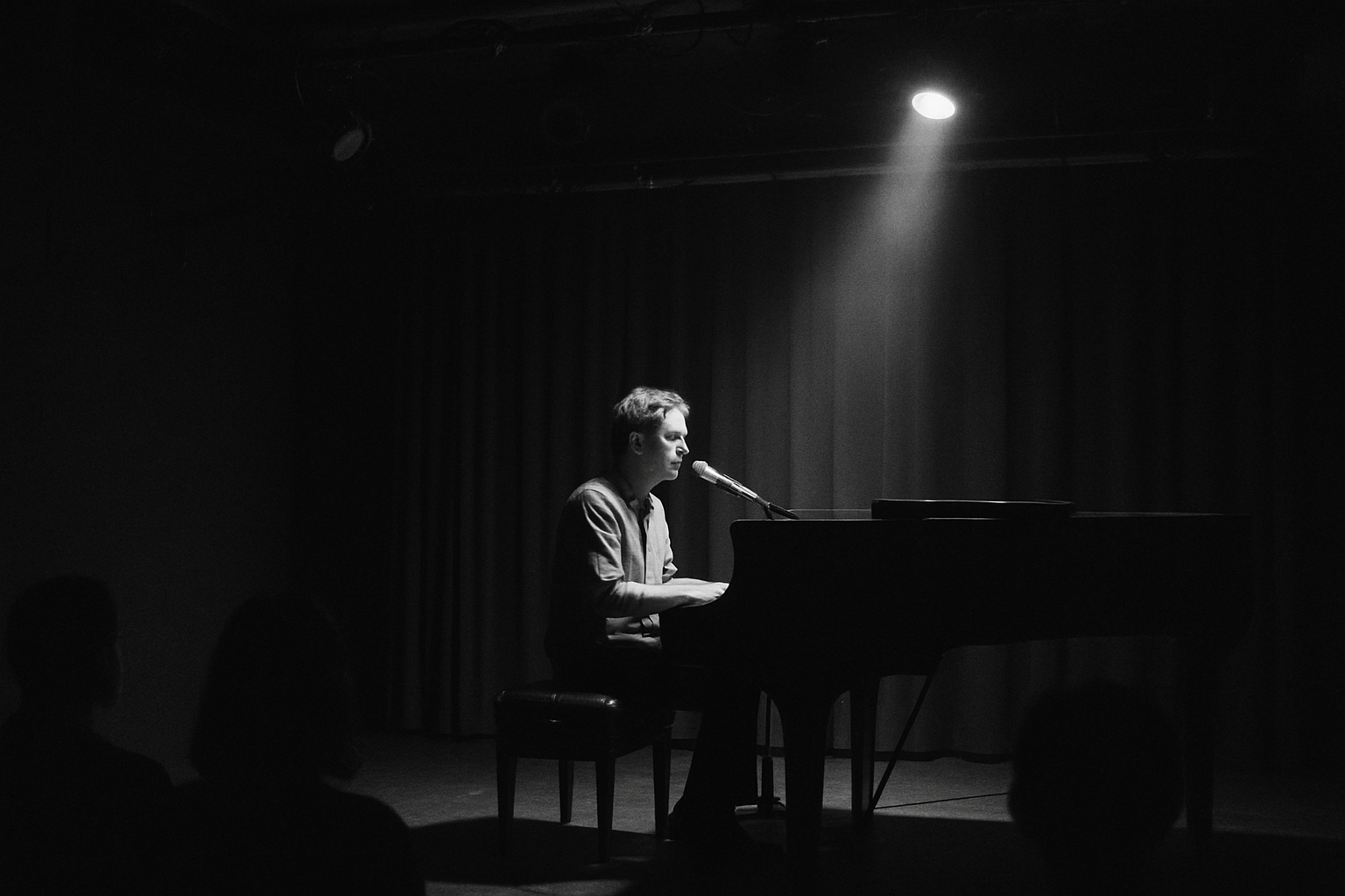 Wide shot of a restrained monochrome stage performance with a songwriter seated at a piano under soft spotlight