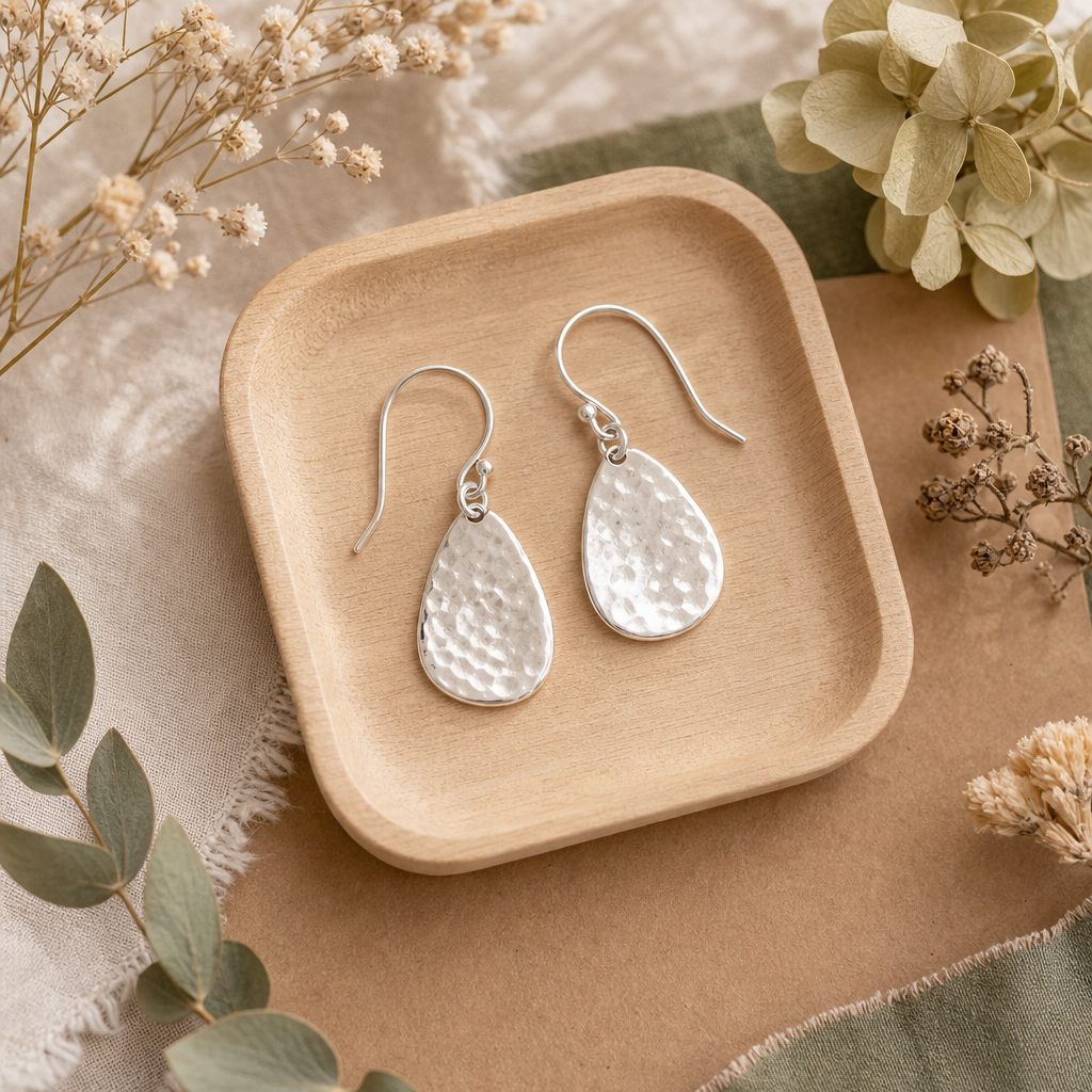 Pair of hammered silver drop earrings arranged on a small wooden tray with dried flowers