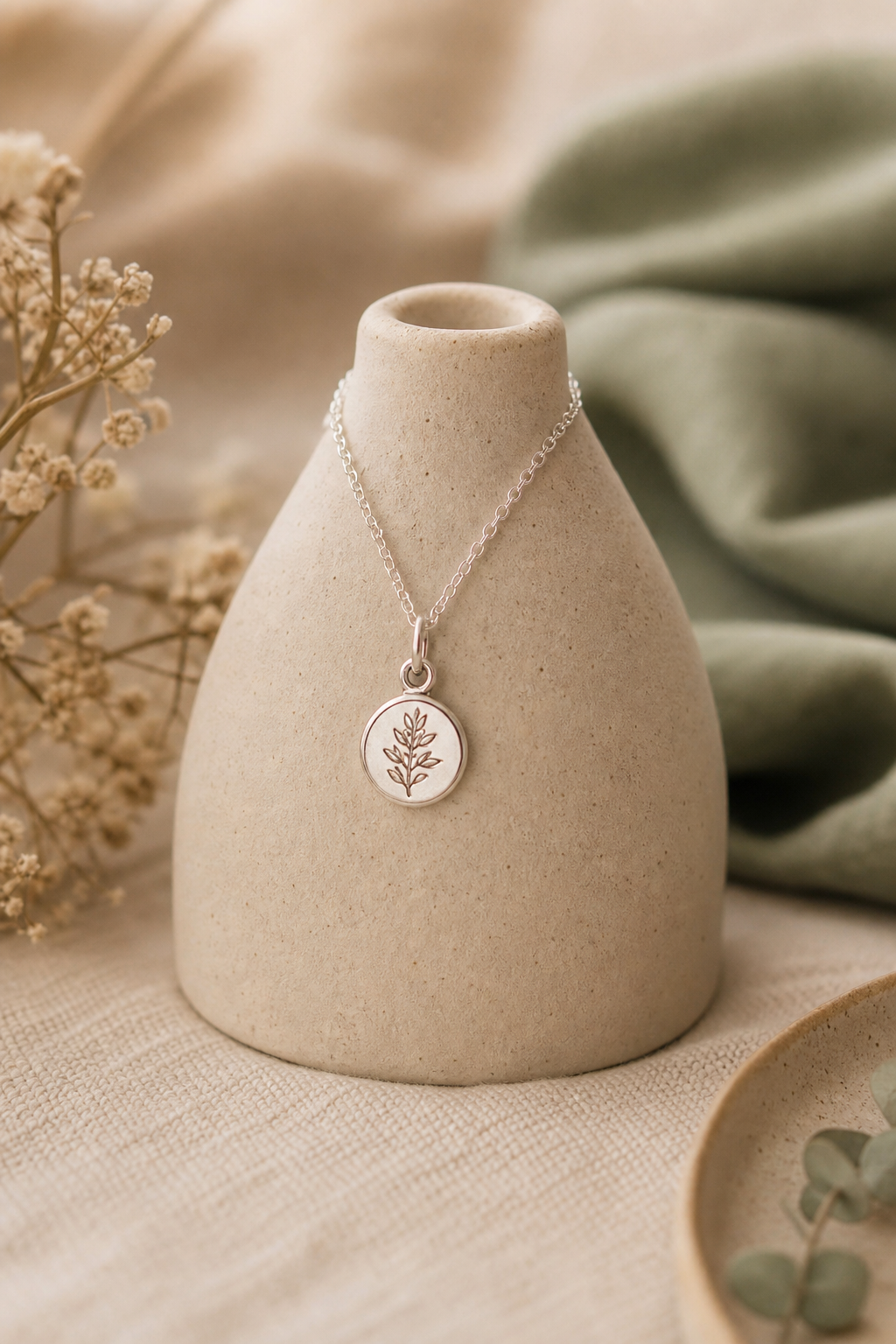 A dainty handcrafted silver pendant necklace draped over a small ceramic stand against a cozy neutral backdrop