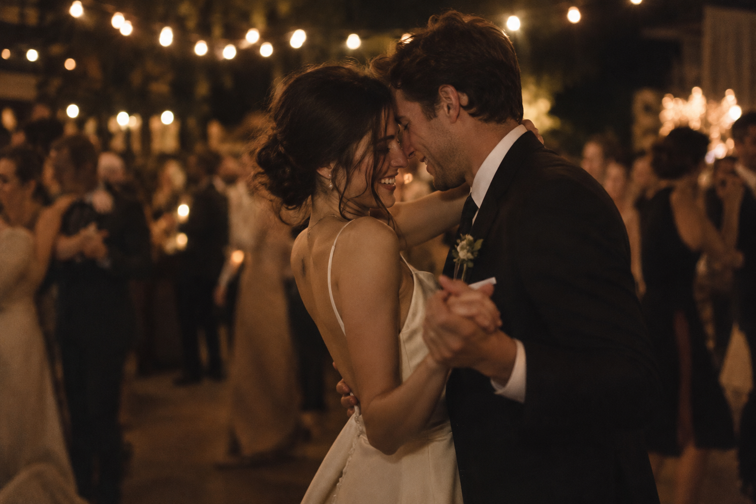 A newlywed couple dancing close together at night, framed by warm reception lights and the movement of their guests.