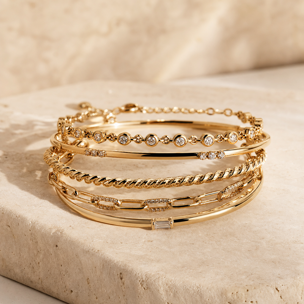 Stack of delicate yellow-gold bracelets with fine detailing on a warm neutral stone