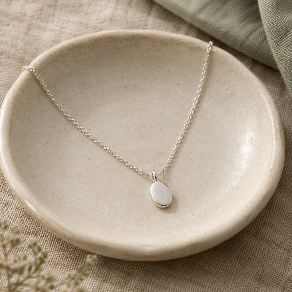A delicate silver necklace with an oval pendant on a cream ceramic dish