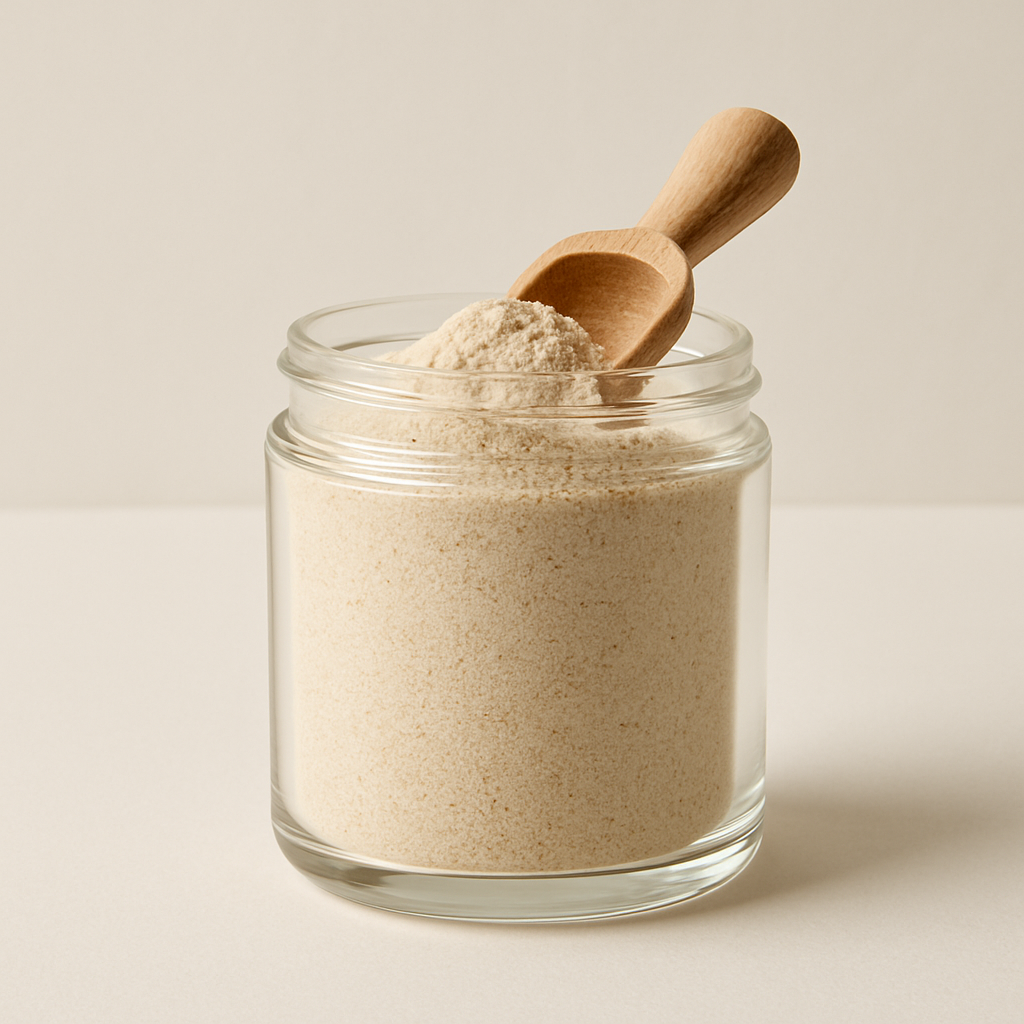 Fine beige supplement powder in a clear glass jar with a small wooden scoop on an off-white surface.