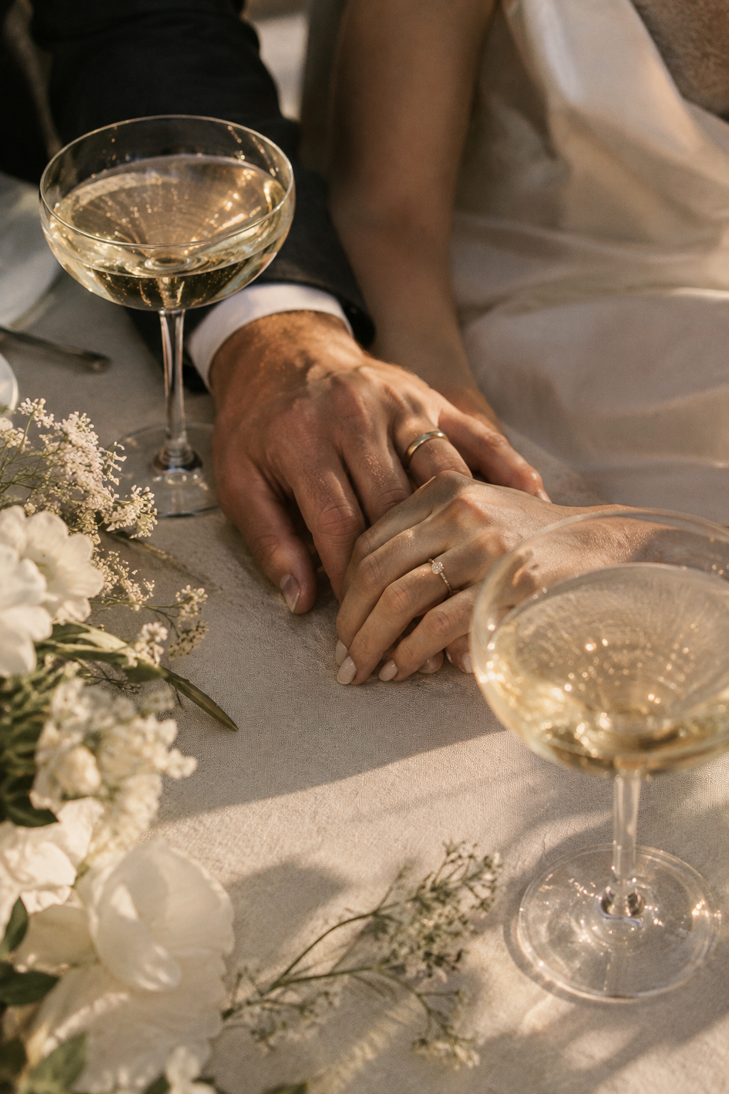 Close-up of champagne glasses and intertwined hands over a linen wedding table in warm evening light