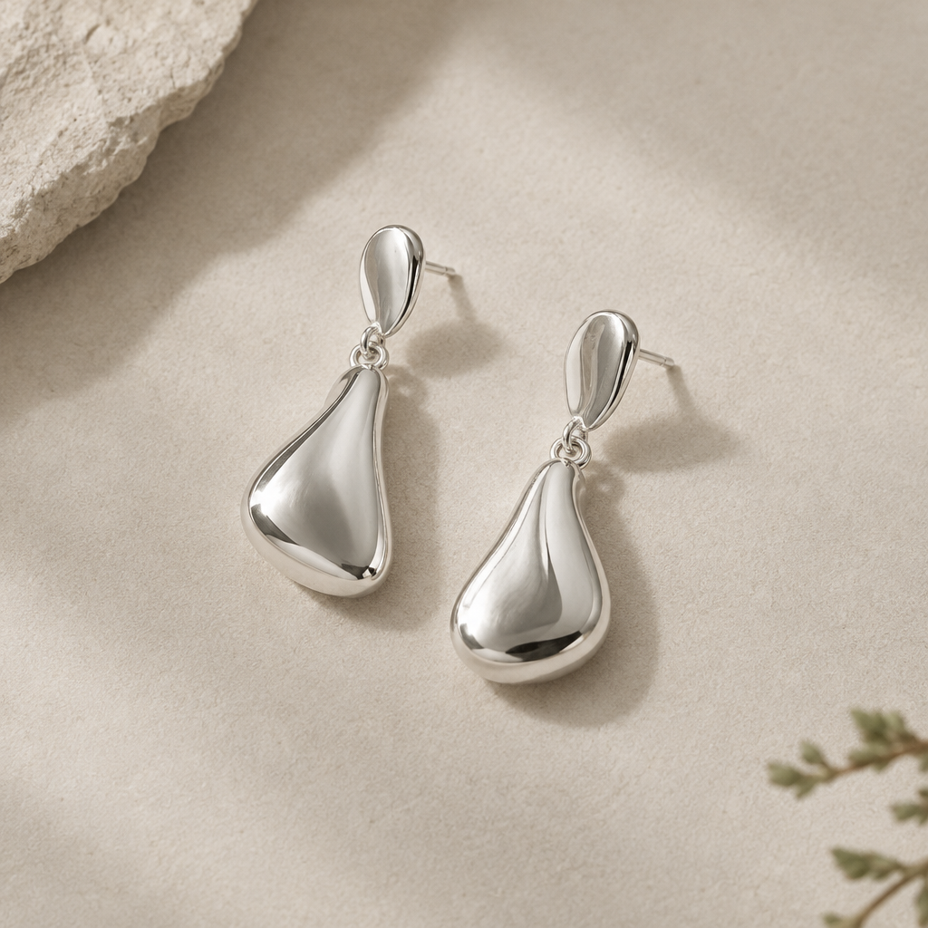Pair of sculptural polished silver drop earrings with softly curved shapes on a pale matte backdrop