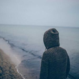 Person in a dark hooded jacket standing on a beach seen from behind