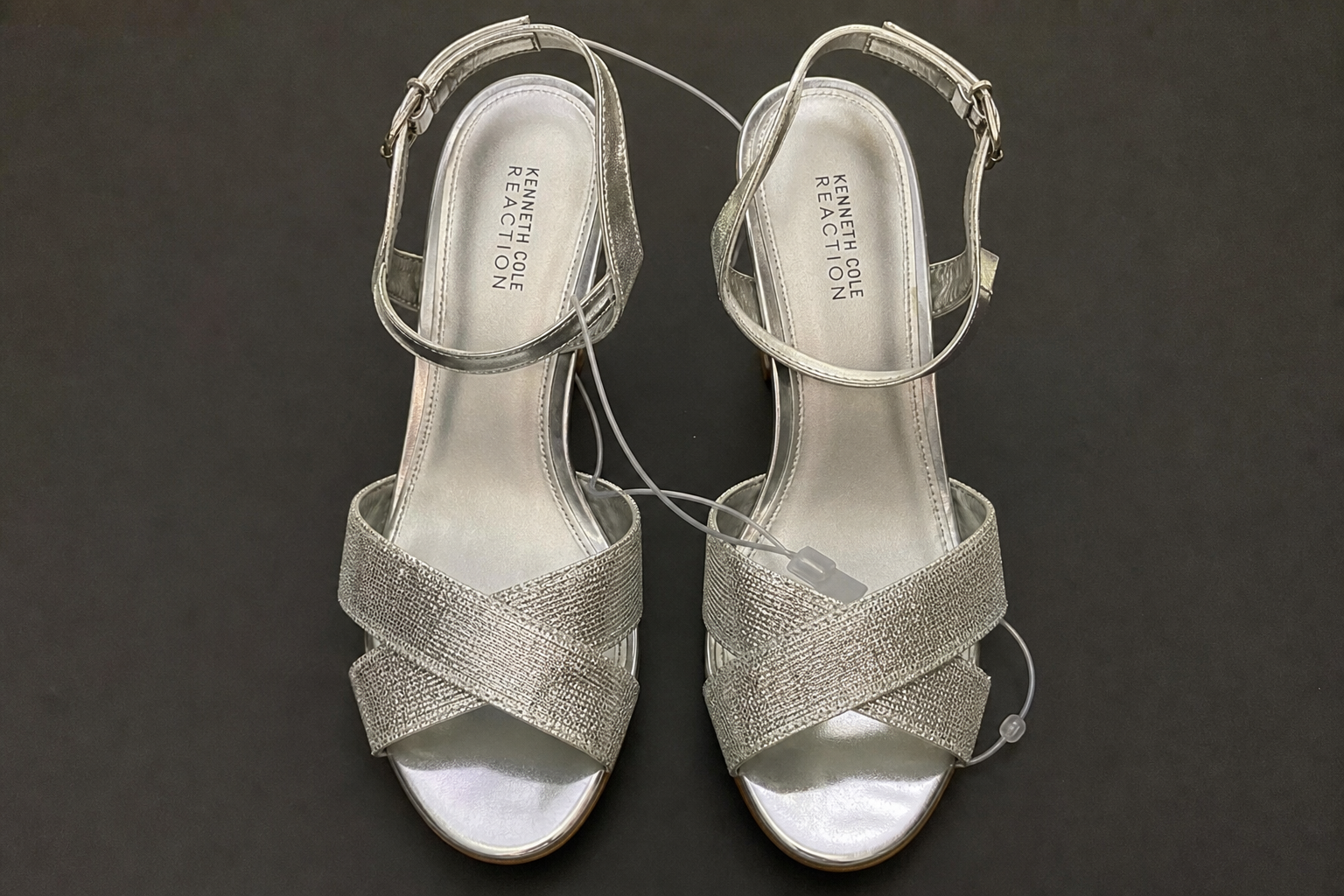 Exact user-uploaded product photo of two Kenneth Cole Reaction Reeva metallic silver open-toe platform heels from a top-down angle, with a clear plastic tie/tag visibly looping between the shoes on a plain dark background