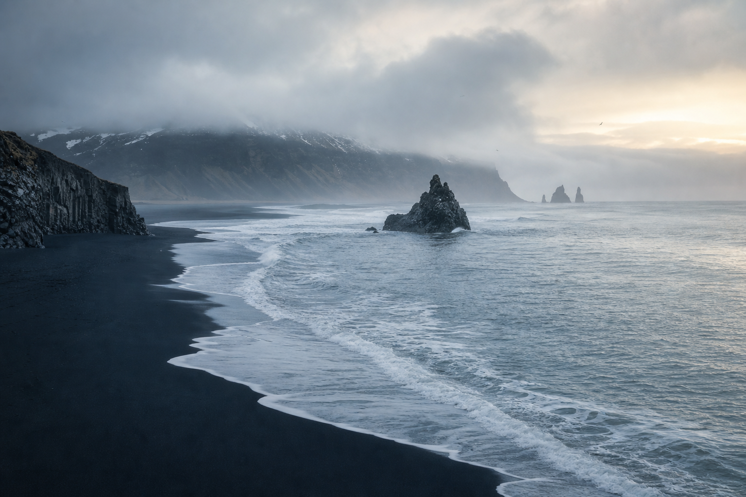 Misty Icelandic coastline with black sand, low clouds, and a calm blue-gray ocean at dawn