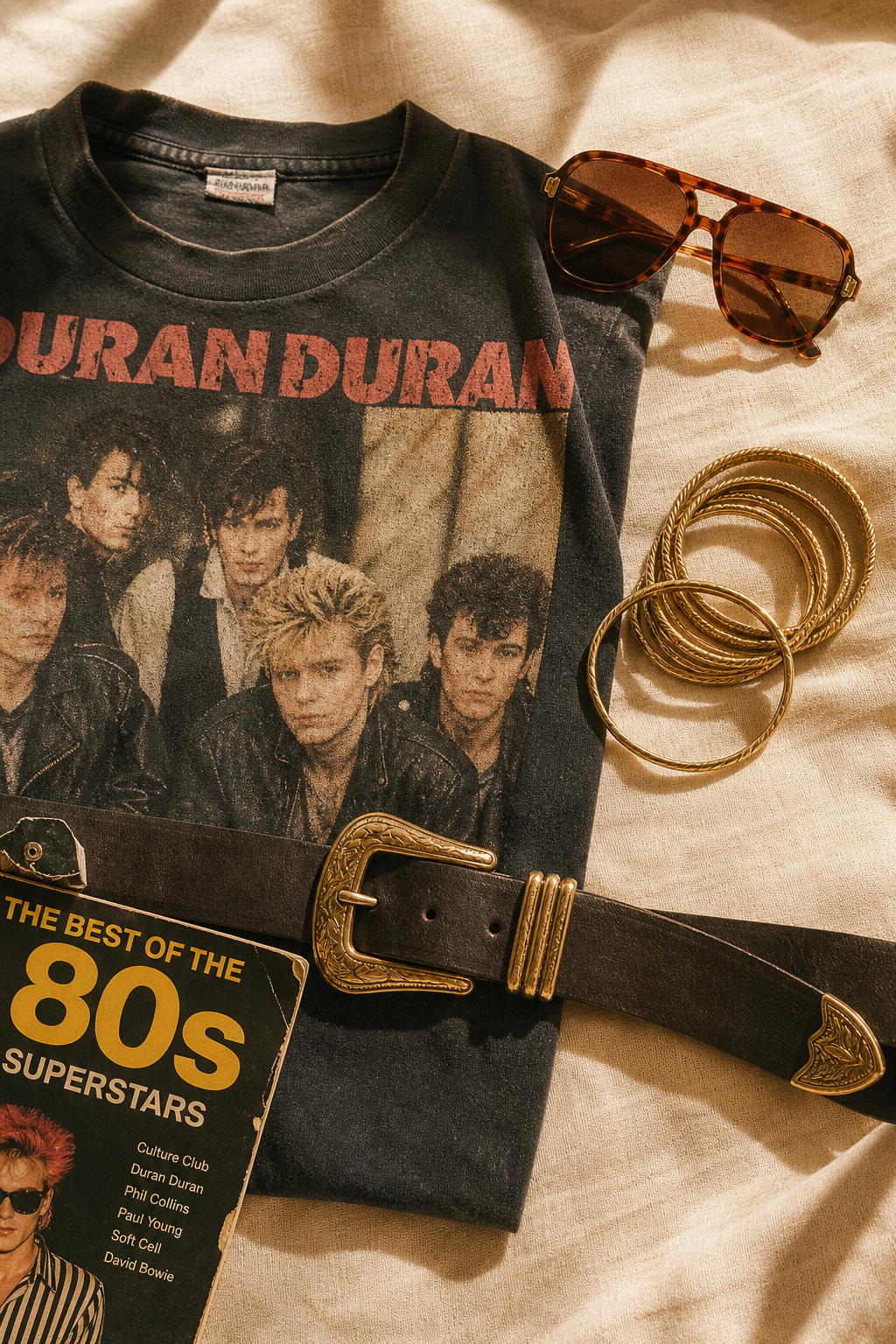 Flatlay of retro 80s pieces including a faded graphic tee, brass belt, and aviator sunglasses on a textured linen surface