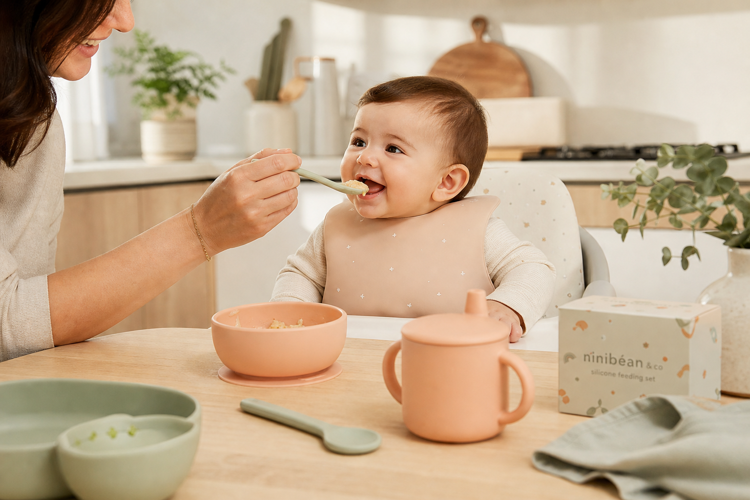 Parent feeding a smiling baby using pastel silicone bowl, cup, and spoon on a light wooden table