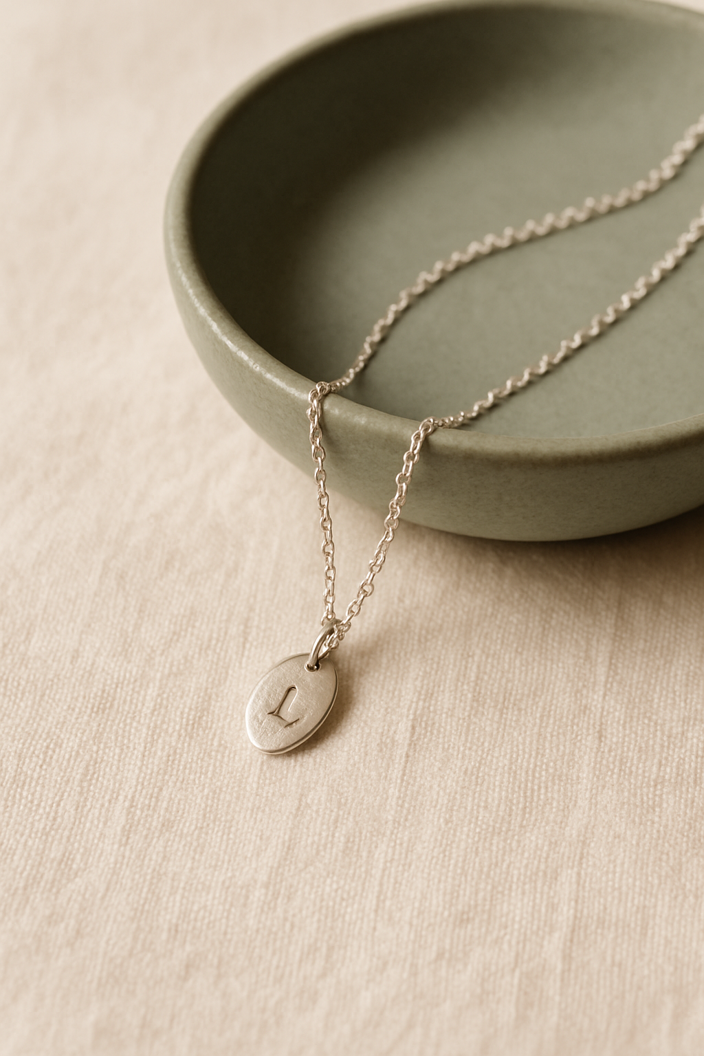 Fine silver chain necklace with a single engraved oval pendant draped over a sage green ceramic dish